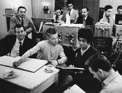 Inside a crowded recording studio a Black woman in a chic black dress and a pearl necklace reads over a musical score with a white man wearing a turtleneck sweater.