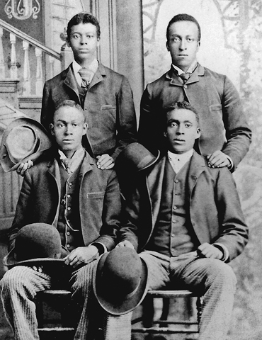 Four young men, their hair combed and parted, are dressed in suits and hold hats. Two are seated, and two stand just behind them, hands casually resting on their brothers’ shoulders.