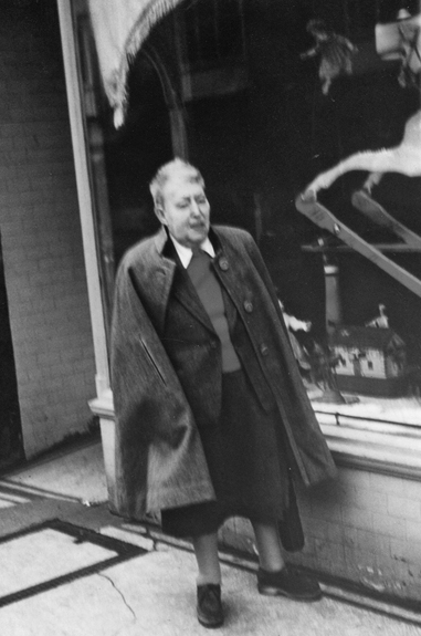 Figure 17. An older Heap with short, grey hair stands in front of a shop window, facing right; her expression is neutral. She wears an unbuttoned cape over a tailored suit jacket and loose, mid-calf skirt. Visible under her jacket is a red sweater over a white, collared shirt.