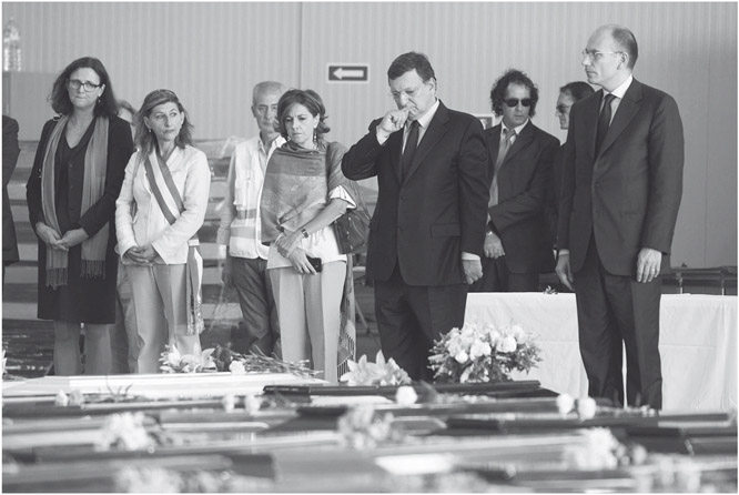 Figure 4. A small group of people stand behind coffins and look down at them with serious expressions. A woman wears the Italian tricolor ribbon across her upper body.