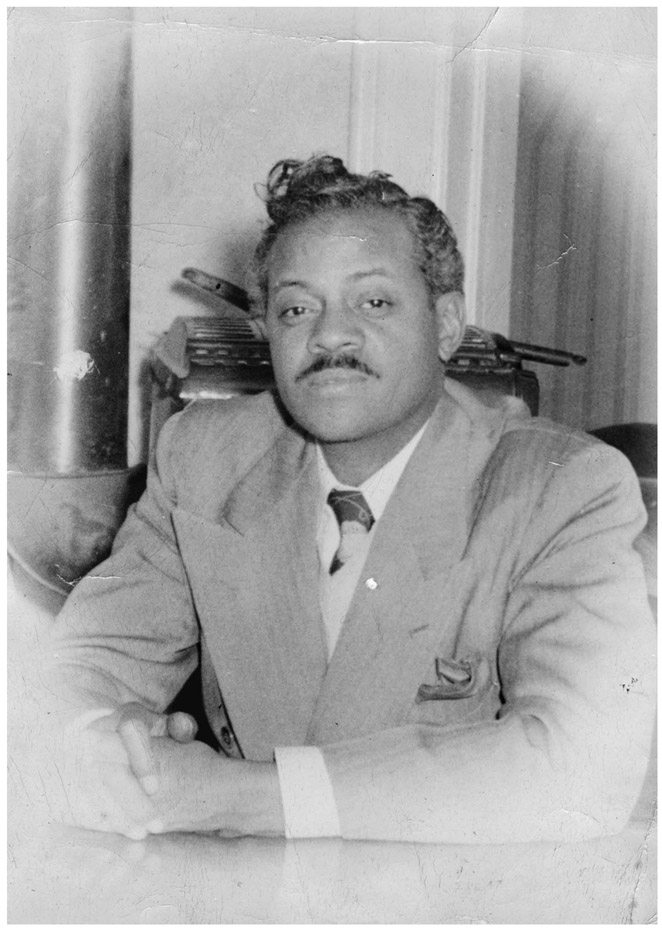A confident-looking man with a thin mustache, carefully combed hair and a well-tailored suit sits in an office in front of a typewriter. The photo is black and white and frayed and faded at the edges.