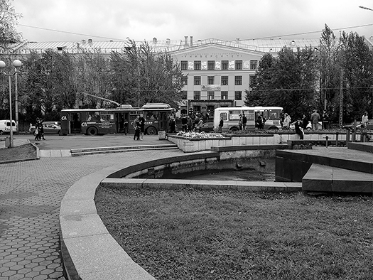 Viewed at a distance across a park plaza, pedestrians wait on a sidewalk for buses and trolleybuses. A pastel painted building in Italian renaissance style and birch trees with early spring leaves are visible across the street. Low light-gray clouds cover the sky.