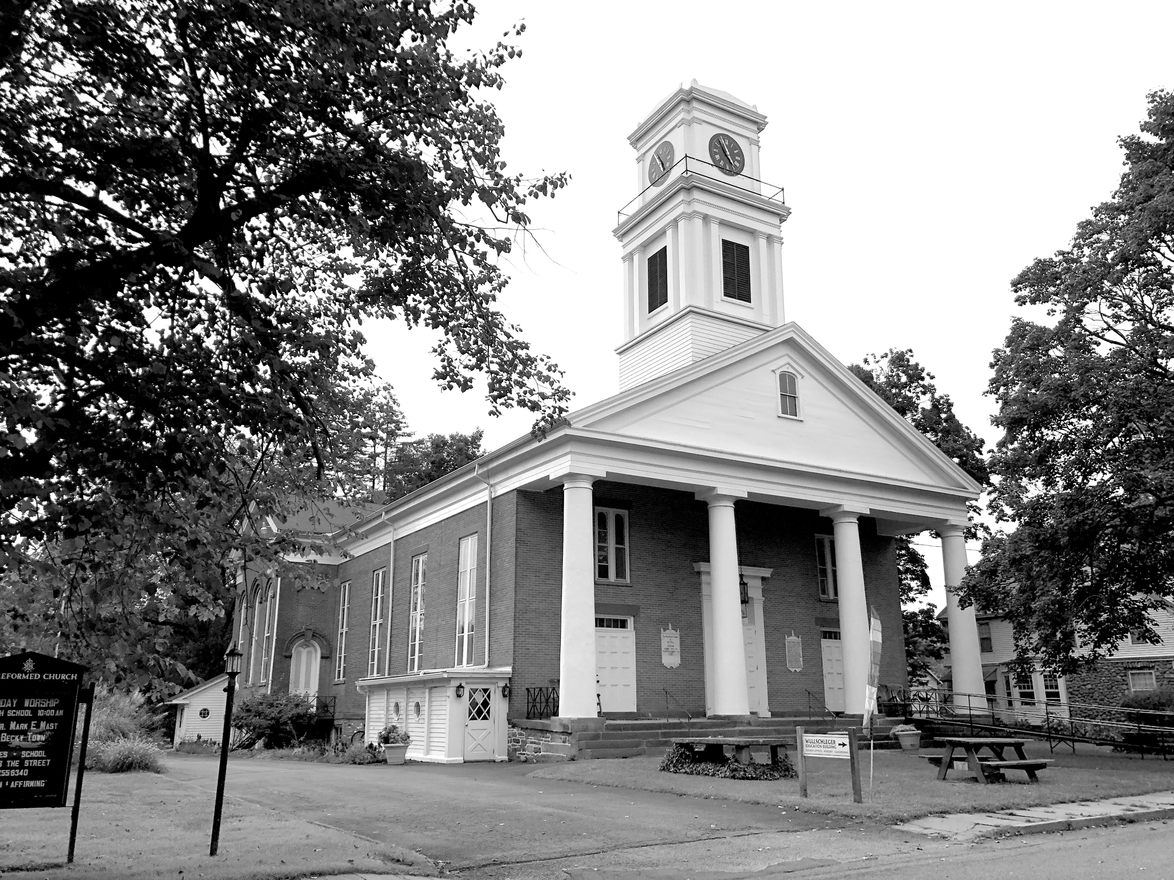 A photograph of a red brick church with white portico and bell tower with clock. The portico has a large white entrance and two smaller side entrances. Picnic tables, a sign, and a rainbow flag are at the front of the church. A driveway on the left of the building.