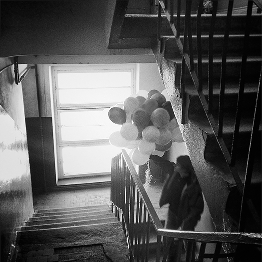The view from the landing in an apartment building staircase shows both descending and ascending flights of stairs. A utility pipe is visible along one wall, and a section of the landing seems to be missing a floor tile. The space is worn but is vibrant and clean.
