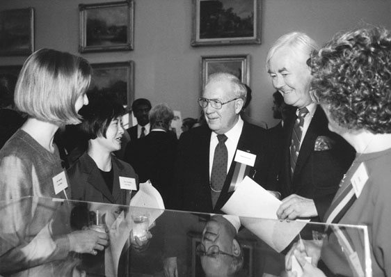 Figure 18 Two young women are holding glasses at a reception while talking with two smiling older men in jackets and ties as a third women looks on. The top of a display case is in the foreground.
