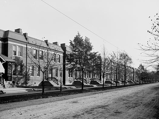 Brick houses in a row showing the front façades and steps, a sidewalk, and trees, with a dirt road in the foreground.