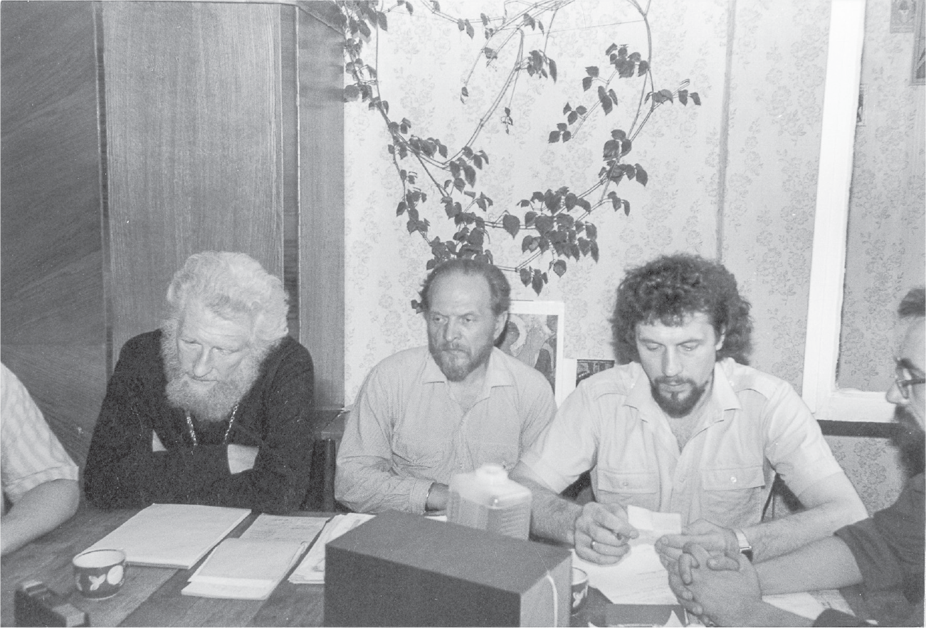 Three men looking serious sit at a table with papers in front of them. The man on the left has thick white hair and full beard, the man in the center has shorter, lighter hair and beard, and the man on the right has curly dark hair and goatee.