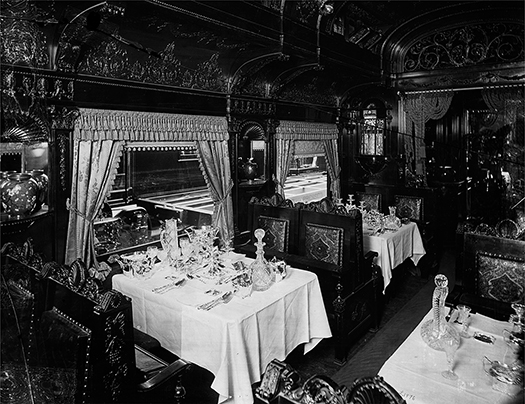 Inside a luxurious railroad passenger car showing tables set with linens and crystal. The walls of the passenger car are carved wood, and curtains adorn the windows.
