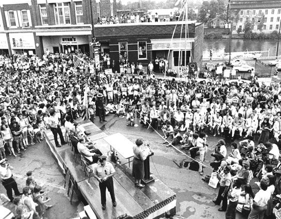 Figure 9 Seen in an overhead view taking in the town’s main street, a dense crowd of onlookers surrounds a raised stage where a woman is speaking. Behind her to one side is a line of dignitaries seated on folding chairs.