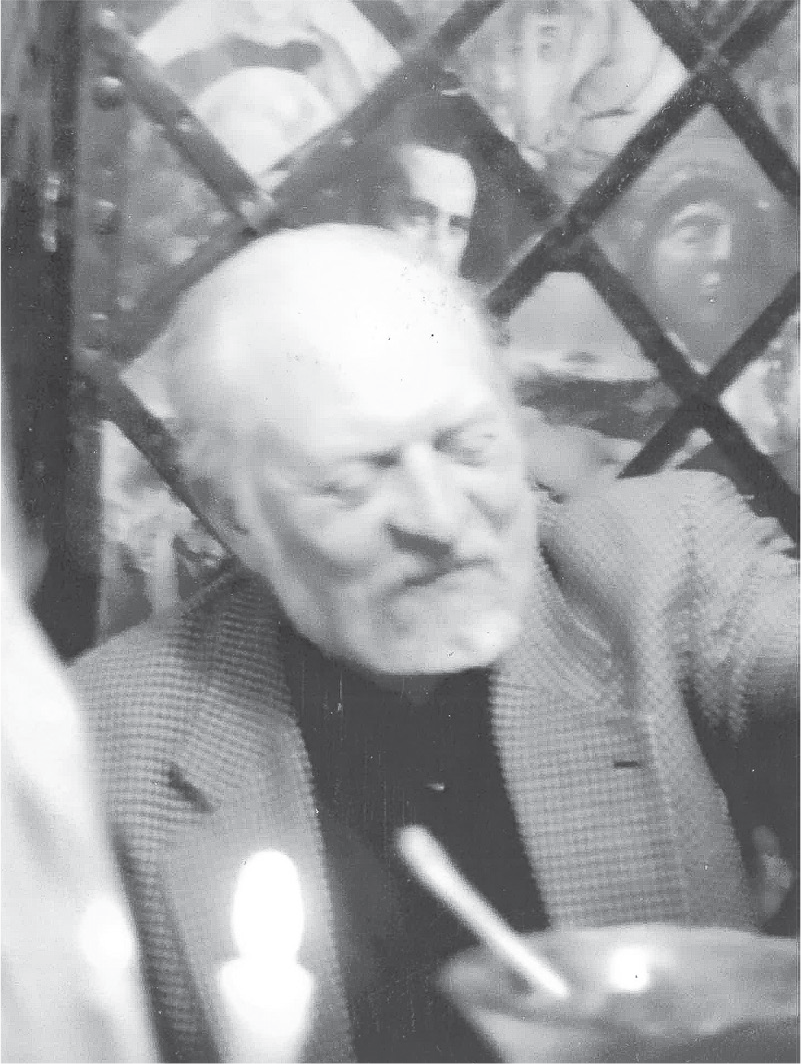 An elderly bearded man in a blazer looks down, smiling slightly. A bowl and utensil are in front of him and behind him is a standing wall of pictures in diamond-shaped frames.