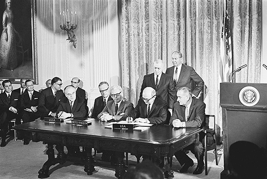 Four men wearing suits sit at a table next to a lectern. One is signing a document. The other men sit and stand behind them.