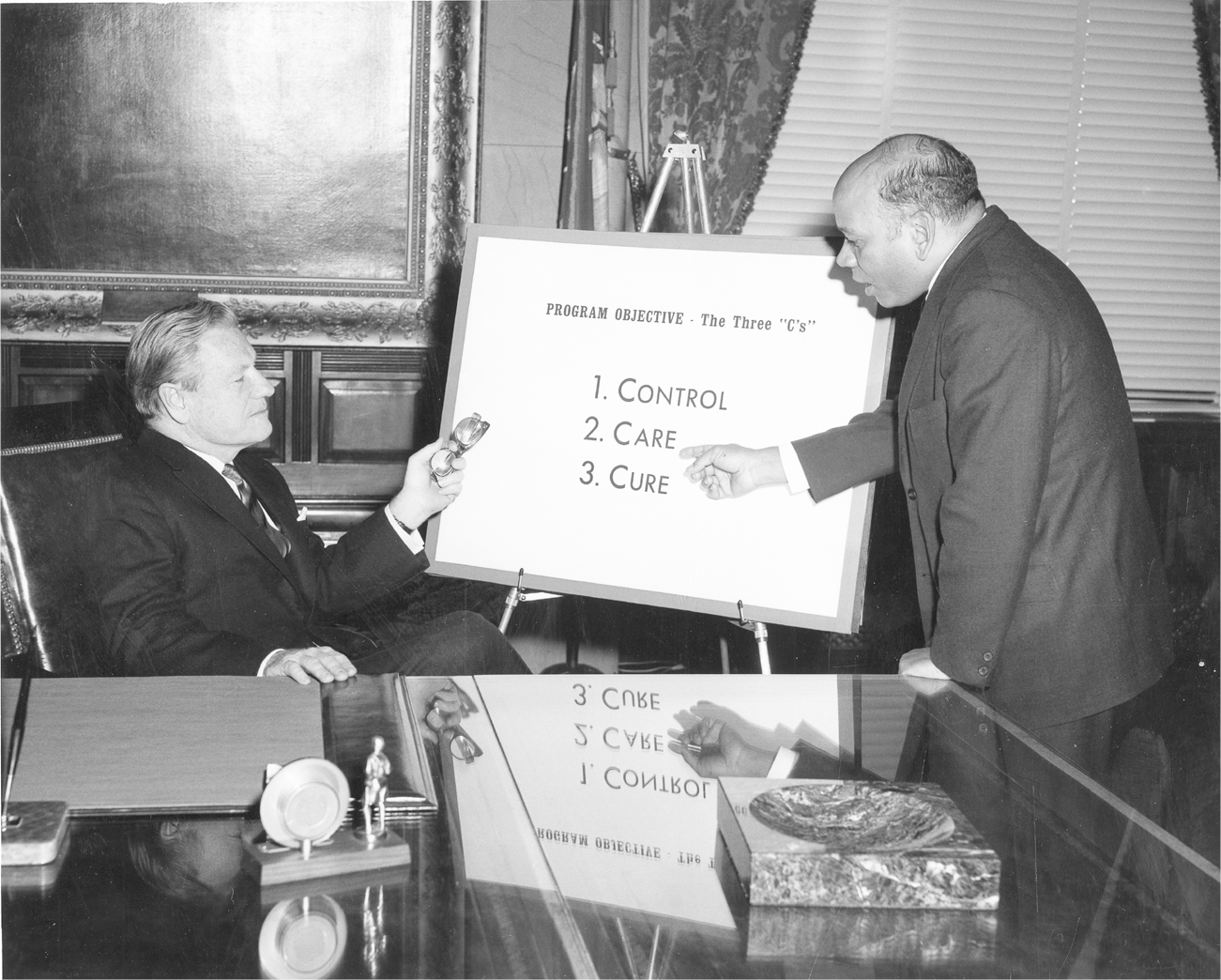Rockefeller sits at a desk in a formal room gesturing toward a sign using his folded glasses as a pointer. A second man stands on the other side of the sign pointing to the words that read, “Program Objective: The Three ‘C’s’ 1. Control, 2. Care, and 3. Cure.”