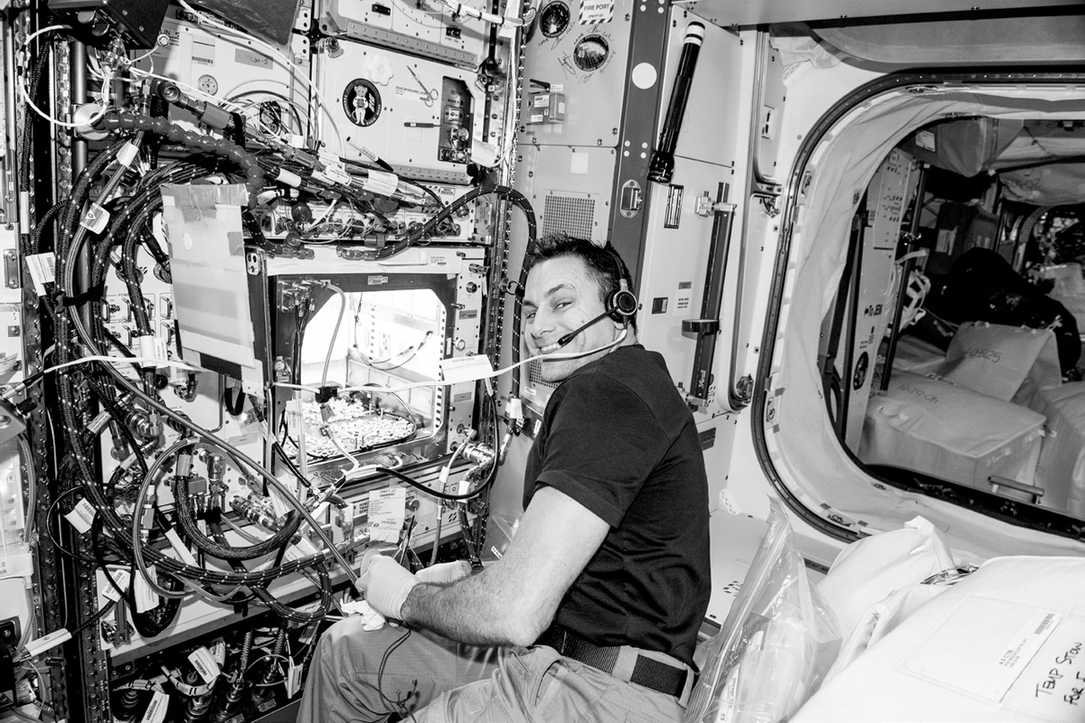 A smiling man wearing a headset sits in front of a large machine covered in wires. There is an opening in the machine through which small seed sprouts can be seen under a bright light.