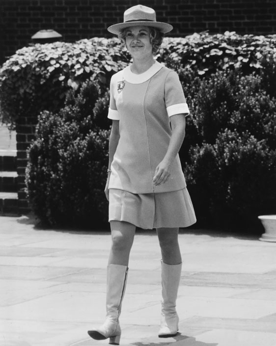 Figure 3. A smiling woman in a mod-looking short-sleeve blouse, matching miniskirt-like culottes, and high white zippered boots and ranger hat strides across a paved plaza.