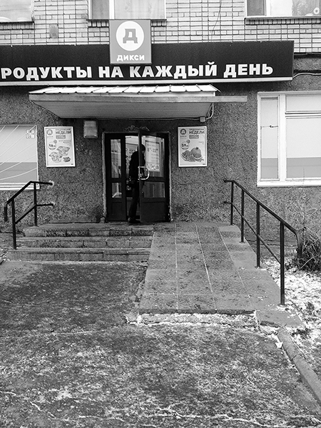 A commercial façade with a sign that says “Groceries for everyday” in Russian. A person enters the shop on foot through a swinging door at the top of a tiled stoop with three steps and a ramp. A light dusting of snow is on the sidewalk.