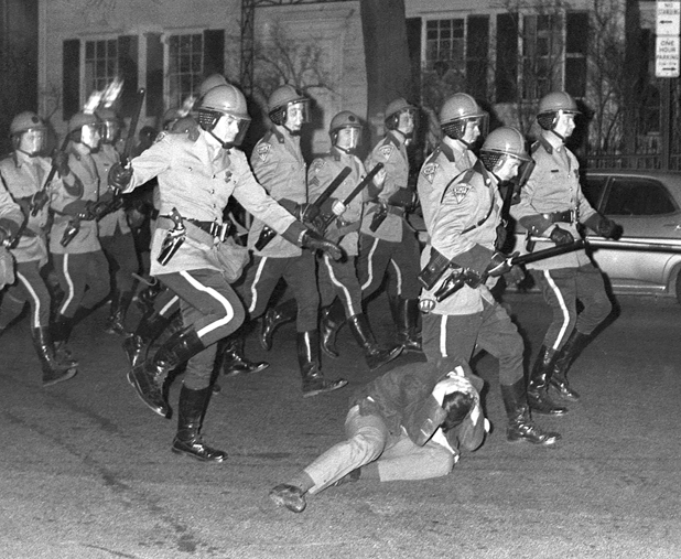 Figure 38. Eleven state troopers are racing down a street, as they pass a fallen young protestor they kick and beat him. He has his hands covering the back of his head attempting to protect himself.