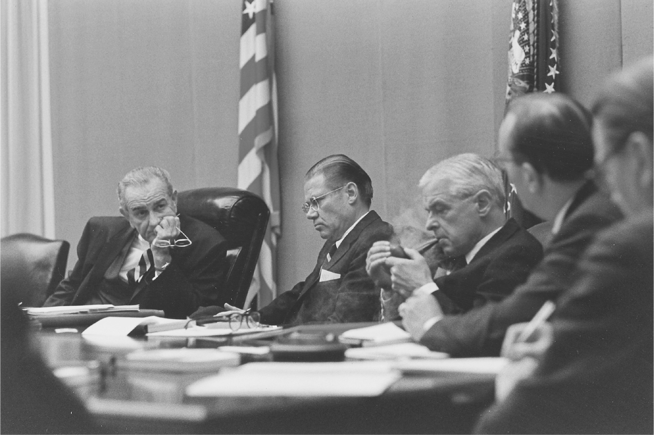 Lyndon Johnson grasps his glasses and holds his hand to his chin while listening to an unidentified adviser. Nitze is lighting a pipe, while McNamara appears to be in a stupor.