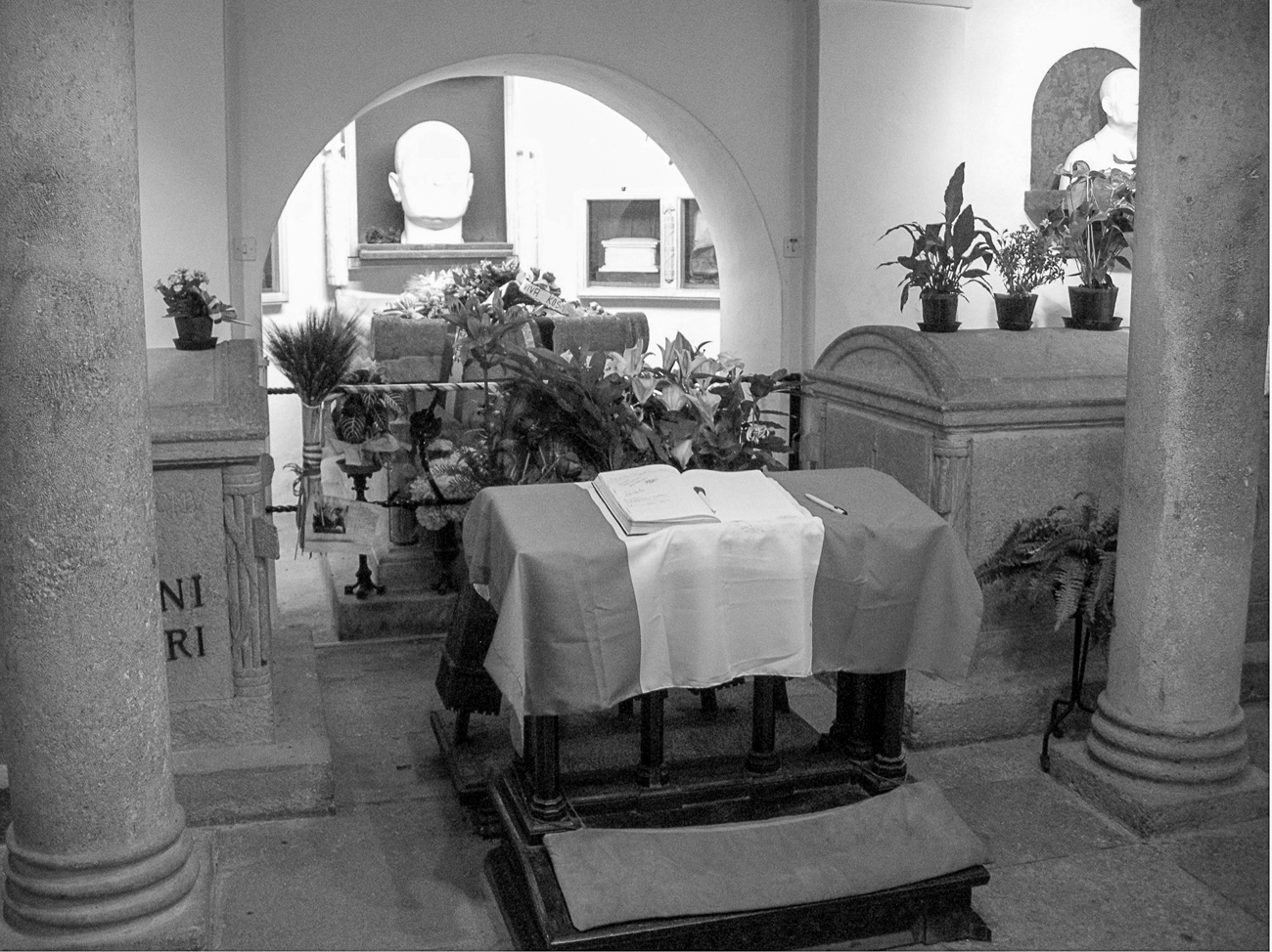 Figure 4.7. A tomb at the back of which sits a large bust of Mussolini. The tomb is decorated by flowers and plants, and at the front sits a desk covered in the Italian flag, with an open book and pen on top of it.