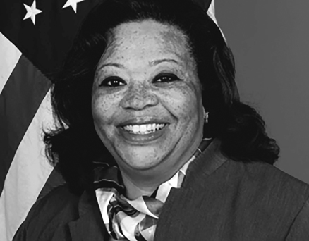 A headshot of a woman with dark, wavy, shoulder-length hair. She is wearing a blazer and a patterned scarf and is smiling broadly. The US flag can be seen in the background over her right shoulder.
