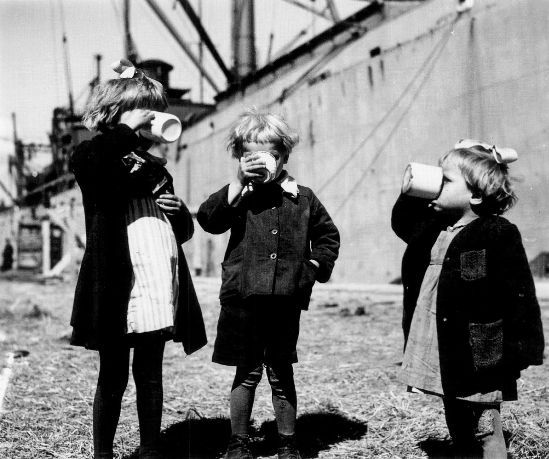 Figure 1. Three small children drink out of mugs while standing in front of a large ship.