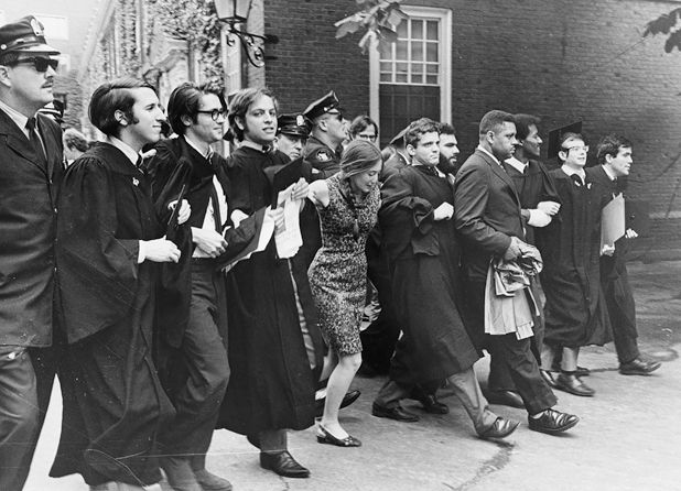 Figure 18. A line of young people, most wearing graduation gowns and carrying mortar boards. All are men except for one young woman in the center wearing a dress, her long hair carefully braided. Behind them are Harvard campus police determined to keep them away from the graduation ceremony.