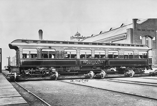 A railroad passenger car being moved by rail on special wheels with a factory in the background.
