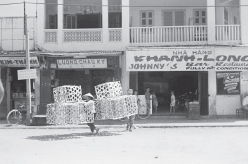 A daytime street scene in Phan Thiet, Vietnam, during the 1960s or 1970s showing two women walking with large crates in front of Johnny’s Bar and Restaurant. The signs are in both English and Vietnamese.