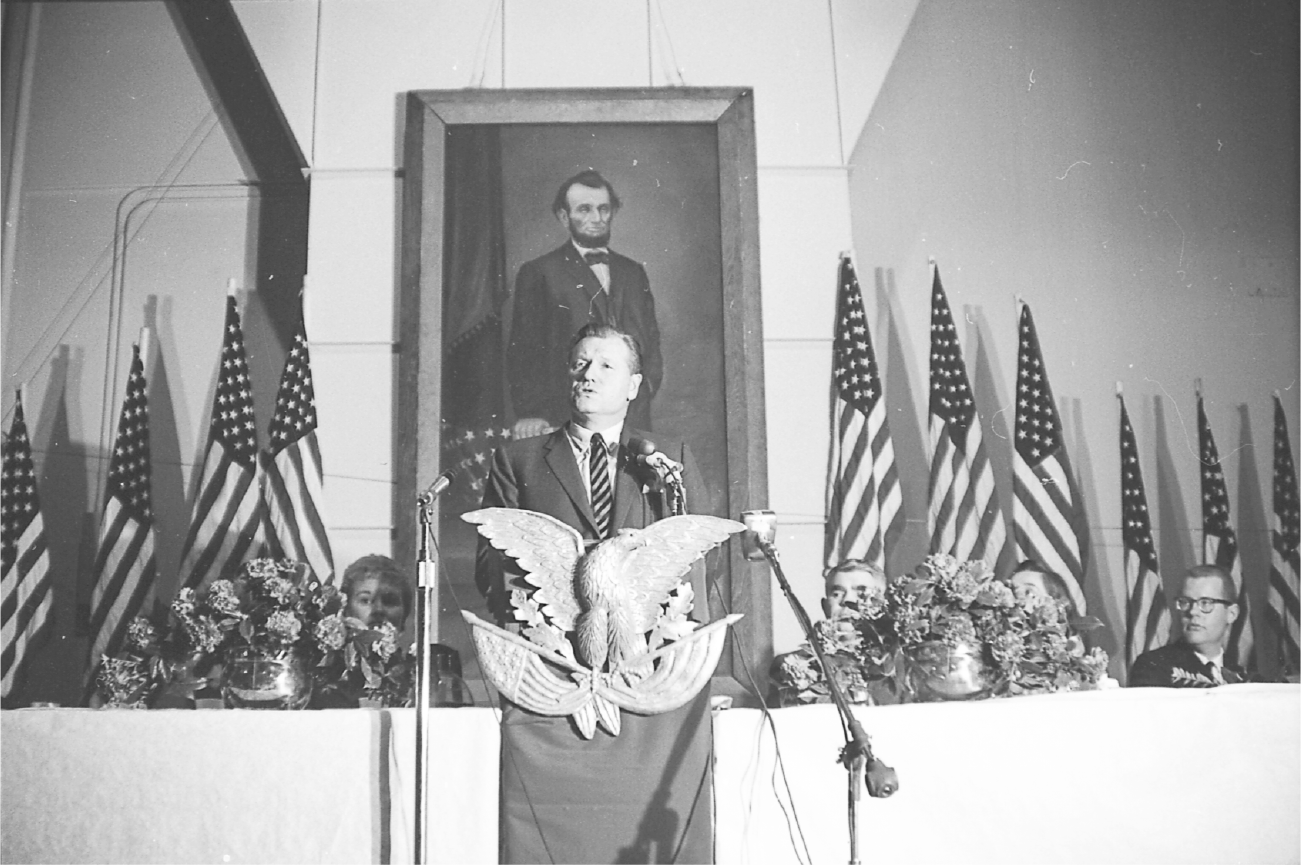 Rockefeller speaks at a podium with a large eagle statuette attached to the front. The podium is flanked by two tables with people sitting to listen to his speech. A painting of President Abraham Lincoln serves as a backdrop.