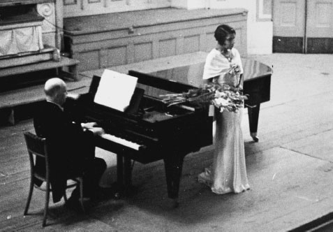 A photograph of a young Black woman in a white dress next to a black grand piano, where a seated man is performing. A bouquet of flowers is perched on the edge of the piano.
