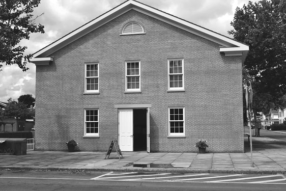 Figure 22 An unadorned two-story brick building has a low-pitched gable front. The symmetrical design includes a central double door with a sash window to either side, with three sash windows spaced evenly above.