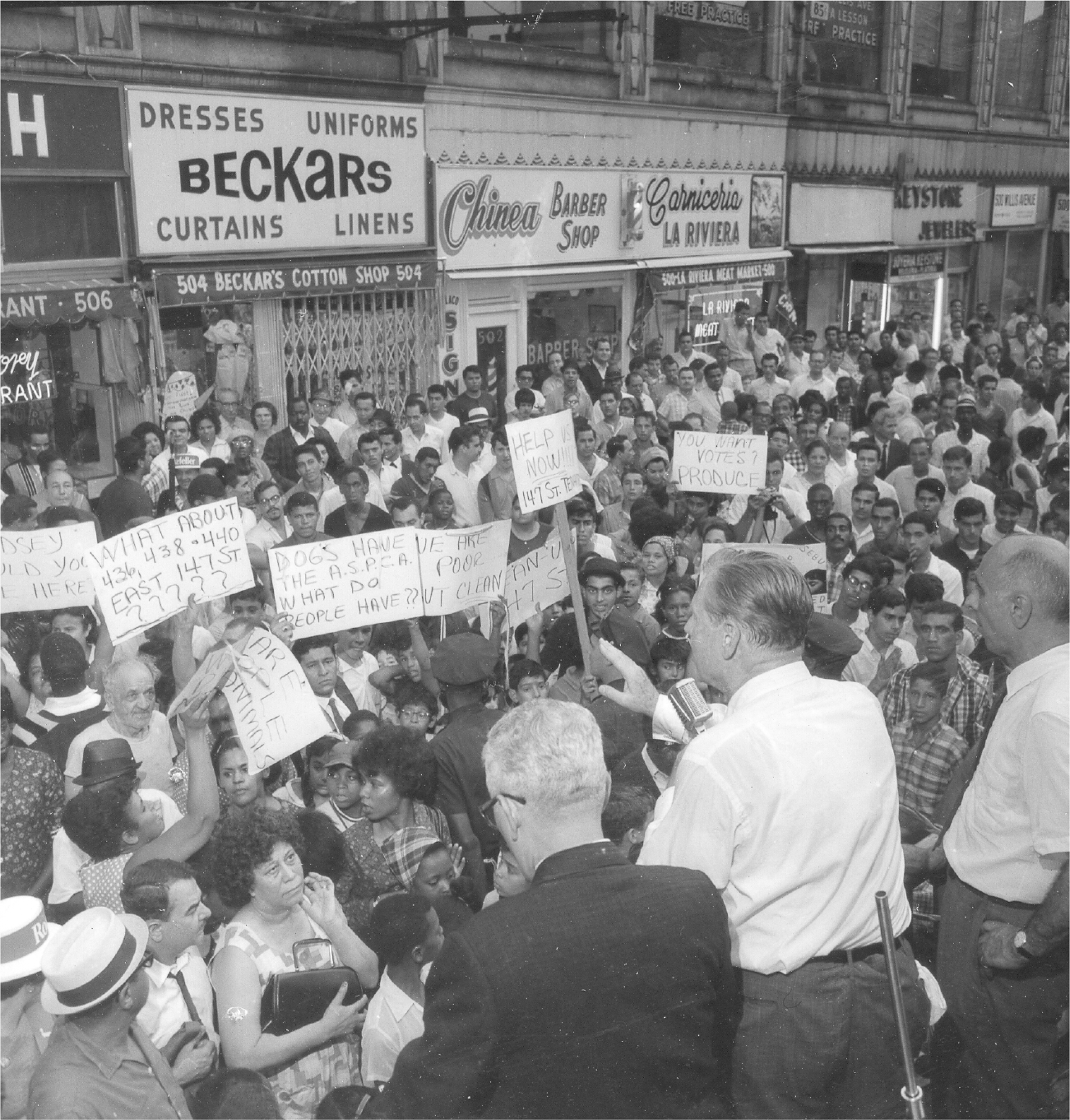 Rockefeller speaks to a large crowd assembled on the street. He uses a microphone as he gestures with his hand. Some people in the crowd hold signs that criticize the governor’s leadership and the state of the neighborhood.