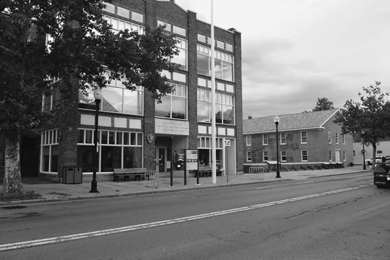 Figure 23 A large, square, symmetrical three-story building of brick and glass has sets of large plate-glass windows across each floor. Park Service signage is visible. The next building down the street to its right is the smaller plain, symmetrical brick Wesleyan Chapel.