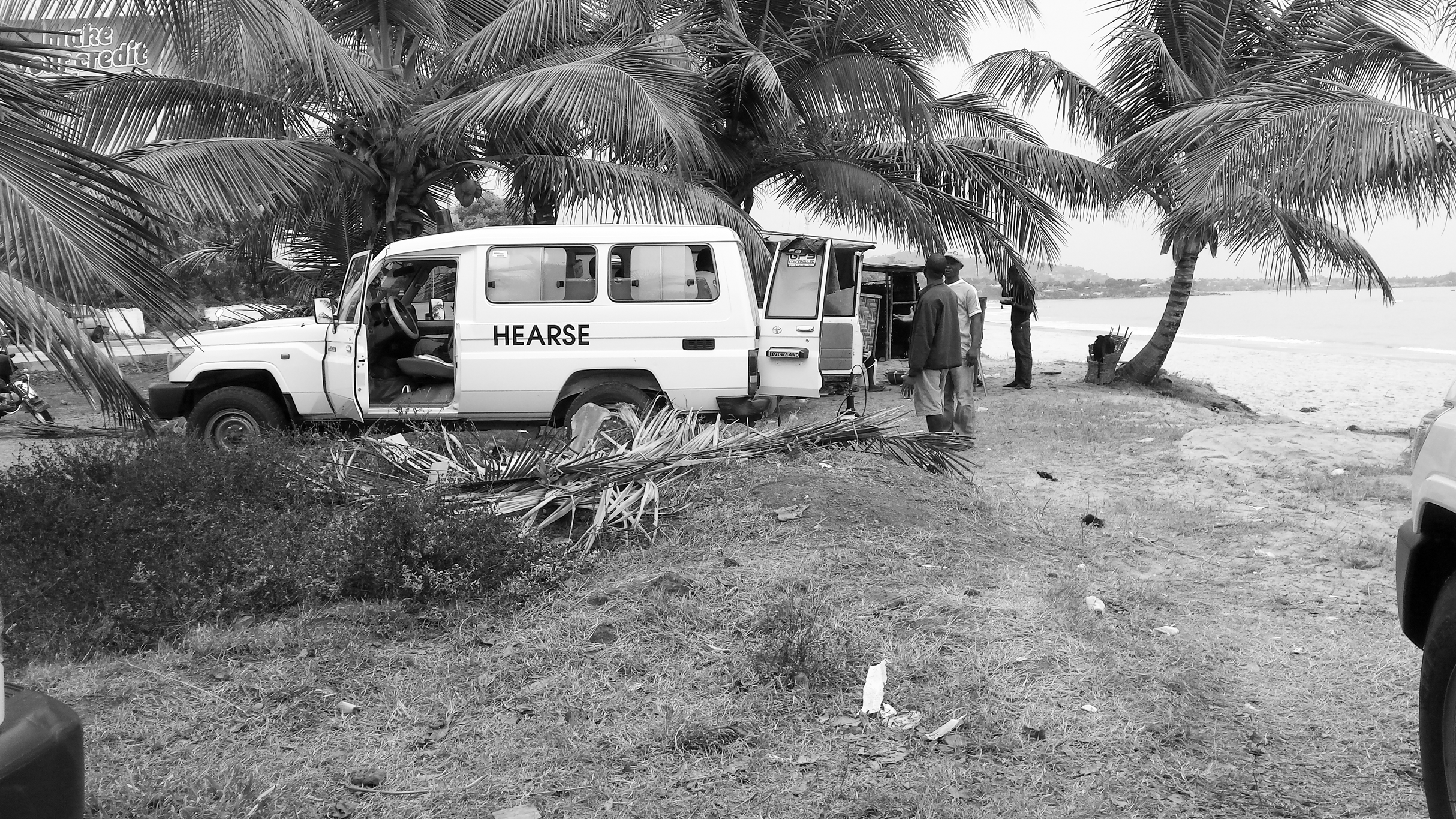 Figure 1. A large white van with “hearse” written on the side is parked with its doors open among palm trees on a beach. Men are standing at the back looking into the van.