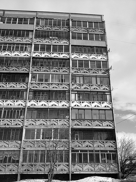 Gray cement panels with triangle designs decorate the exterior façade of an apartment building with identically sized apartments stacked nine high and three across. Apartments on the first two floors have bars on the windows (a safety precaution). A few have replaced the balcony façade with corrugated zinc panels. The sky behind the building has white clouds.