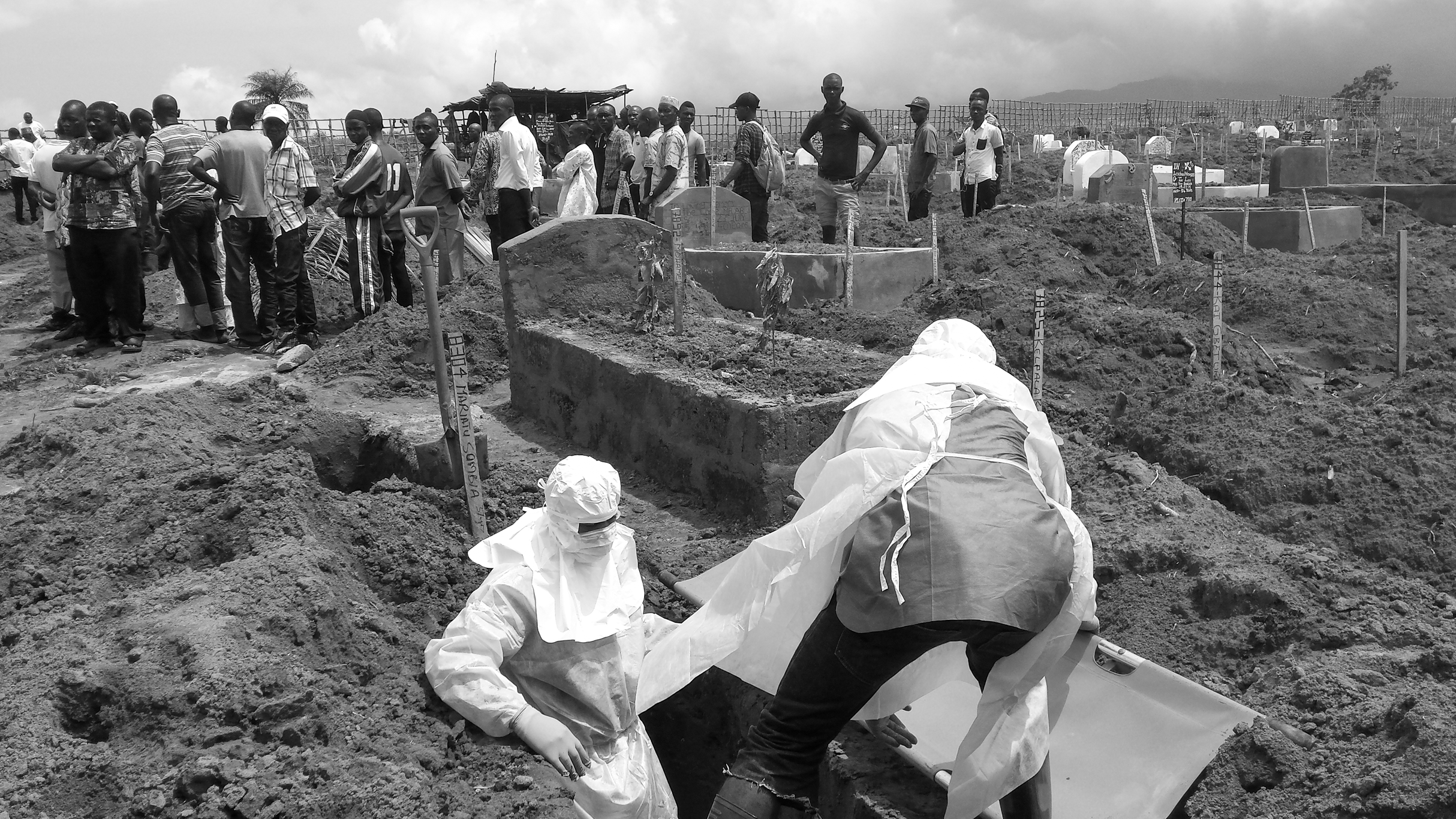 Figure 4. Two people in hazmat suits perform a burial as mourners stand in the background.