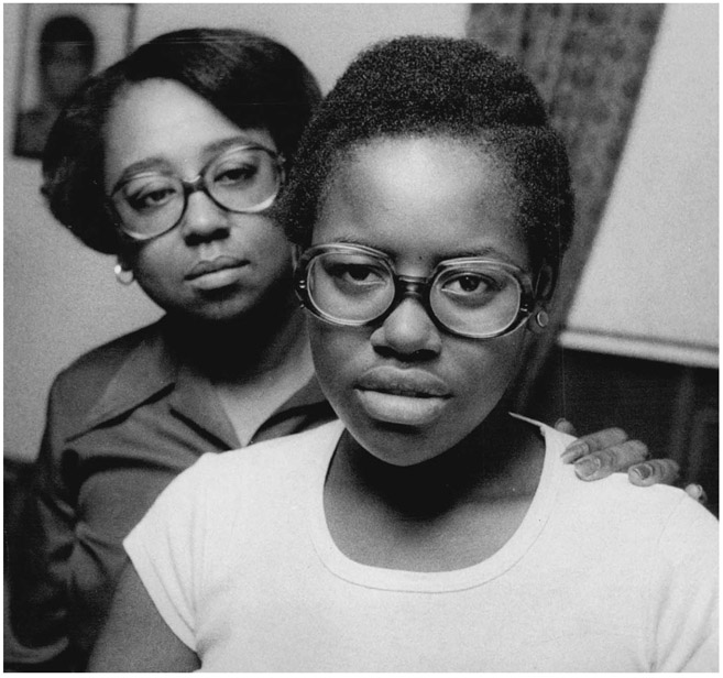 A young Black girl in a white shirt and large round eyeglasses looks intently into the camera. Her mother stands behind her with a look of concern and her left hand on her daughter’s left shoulder.