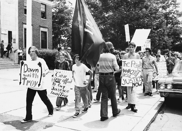 Figure 42. A picket line of young people marching in front of the courthouse with a big flag that says “TPF” and signs that say, “Down with Troy,” “The people want real justice now,” and “Can you afford to buy justice from Troy?”