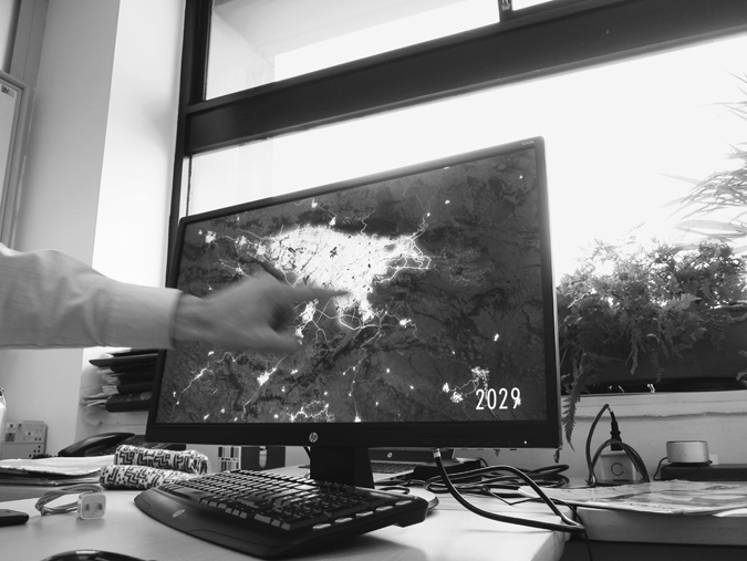 Figure 5.5. A computer monitor sits on a desk in front of a window, and a hand points to a map on it. In the bottom right corner of the computer screen, the text reads “2029.”