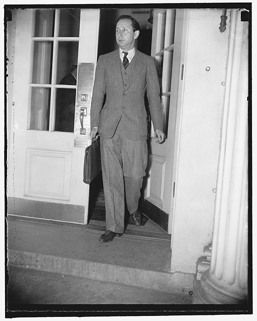 A slim man wearing a suit and carrying a briefcase walks through double doors out of a building.
