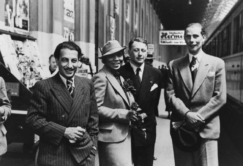 A fashionably-dressed Black woman stands on a train station platform with three men wearing suits.