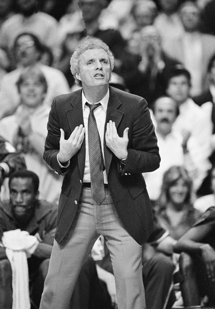 Knicks head coach Hubie Brown stands courtside in a slightly hunched posture, gesturing with his hands and debating a call. Trent Tucker is visible on the bench behind him.