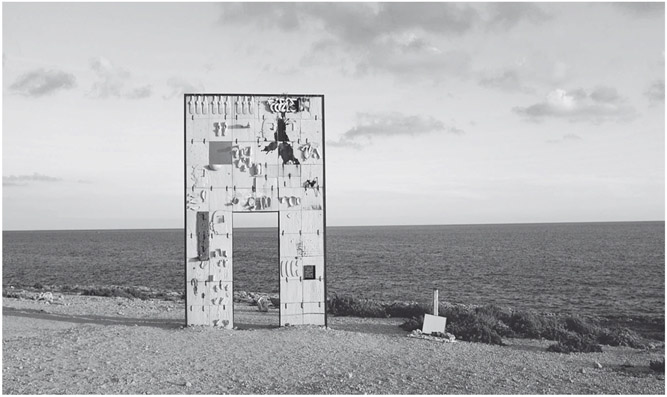 Figure 13. A tall monument resembling a gate stands on a barren terrain close to the seashore. Objects such as cups and shoes made of the same clay material as the surface of the gate are attached to the monument.