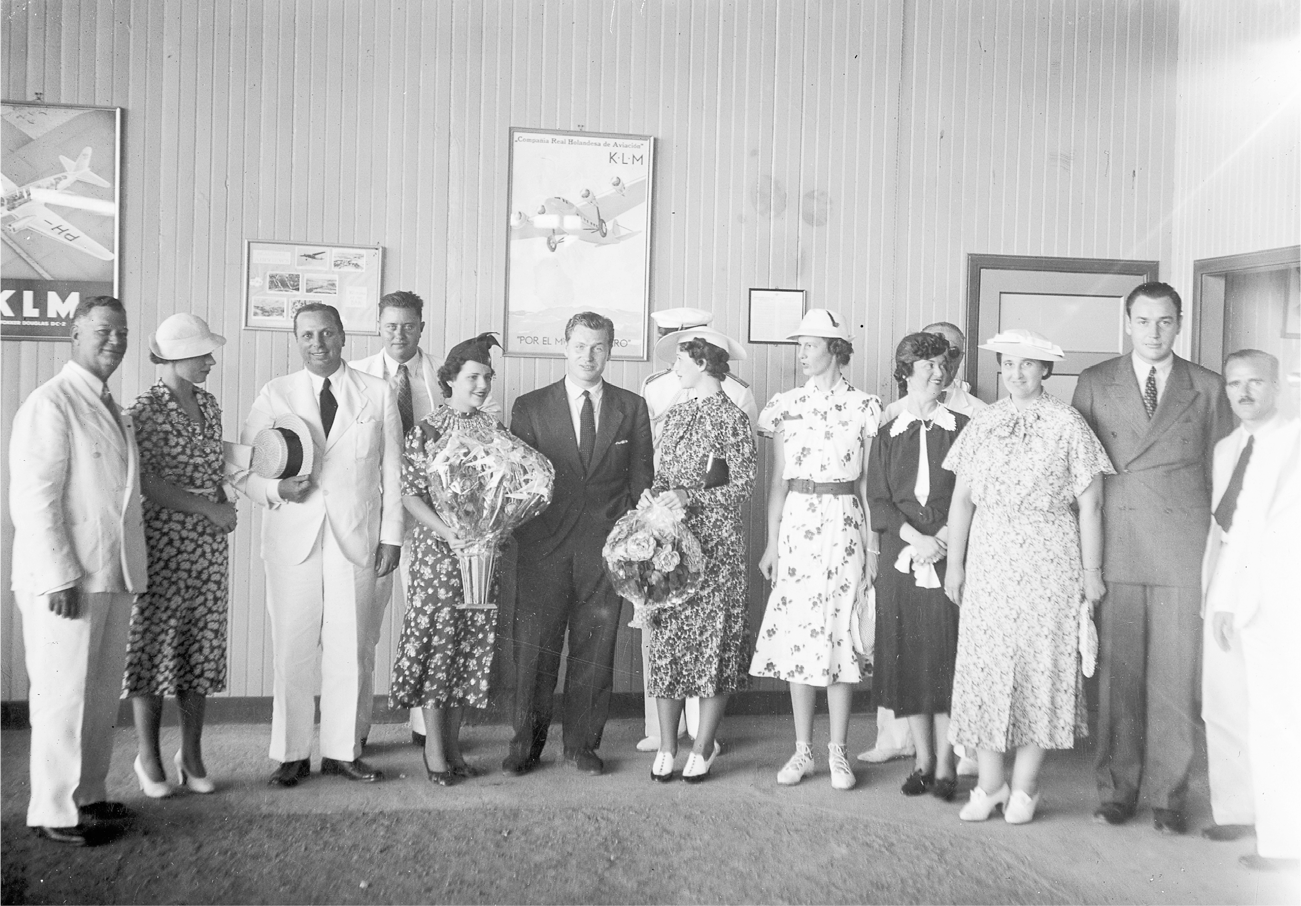 A group of people in relatively formal attire assemble for a photograph in front of travel posters. Nelson Rockefeller is in the center looking directly at the camera while some of the others look in different directions.