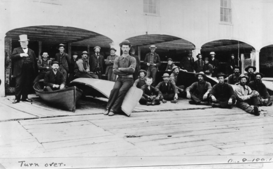 Over twenty-five men, dressed in loose shirts, work pants, vests, and suspenders, sit and stand around their boats in front of a boathouse.