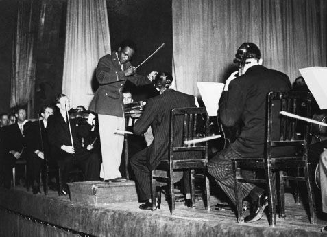 A Black man wearing a US military uniform stands on a short podium holding an orchestra baton in his right hand, leading several dozen men in a musical performance.