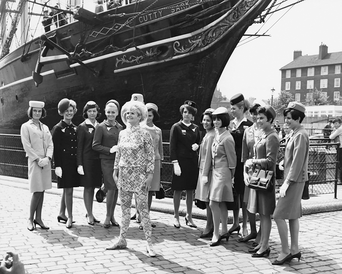 Figure 7.1. A group of about a dozen flight attendants stand in a semicircle around another flight attendant who is dressed in a patterned dress and tights and wearing a bubble cap. A ship and a three-story building are visible behind the women.