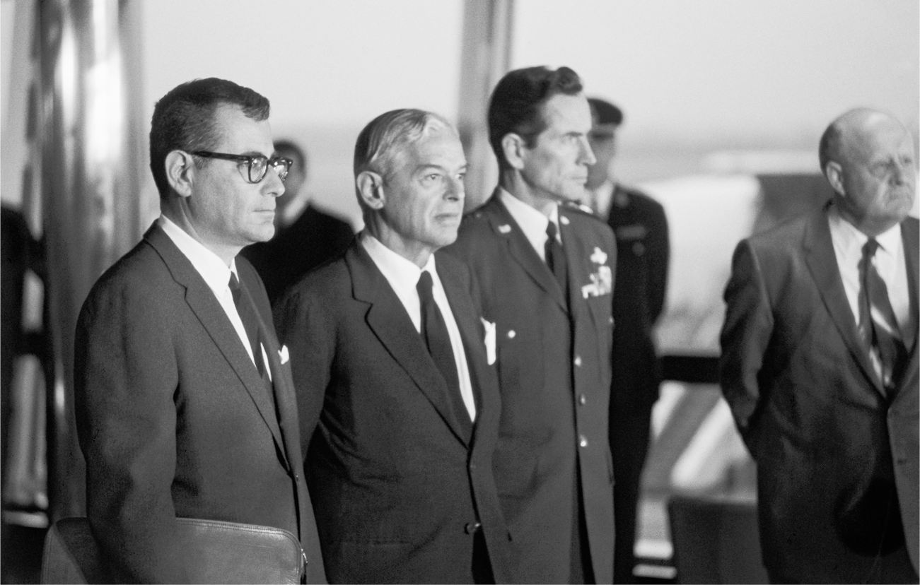 Brown, Nitze, and Allison—in military uniform—stand inside an airport and listen intently to a speaker to the right of the camera.