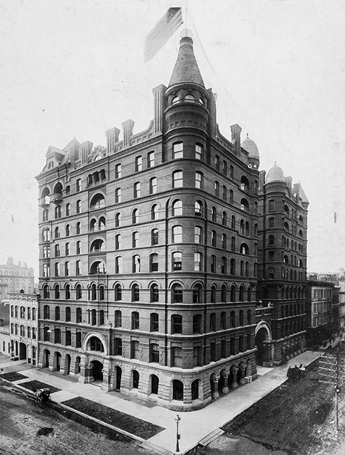 A multi-story brick building on a street corner with sidewalks and roads in the foreground and an American flag on top of the building's roof tower.