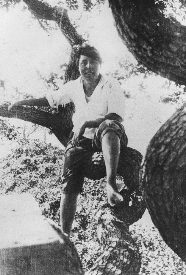 Figure 8. Heap sits casually in a tree, smiling. She is barefoot and has her pants rolled up to her knees. Her right leg is bent, knee forward, with her foot propped on the bough while her left leg hangs over the edge.