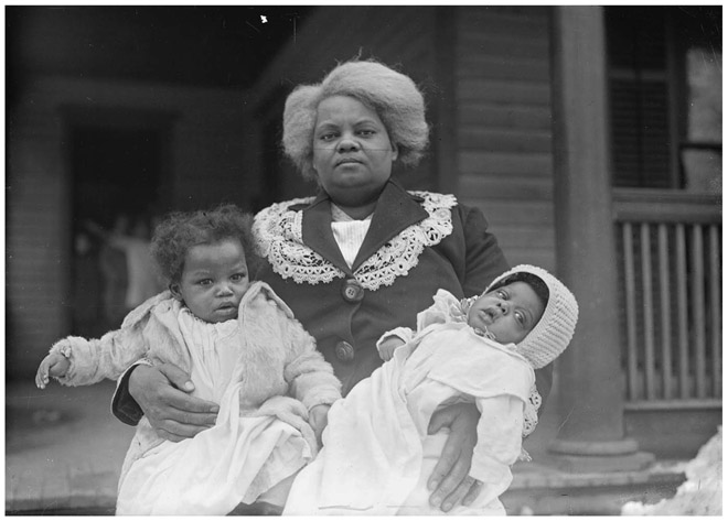 A Black woman with a determined look holds two babies, one in each arm, standing in front of a building where other children are visible in the doorway.